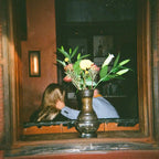 Person sitting at a table with a vase of flowers in front of them, reflected in a mirror.